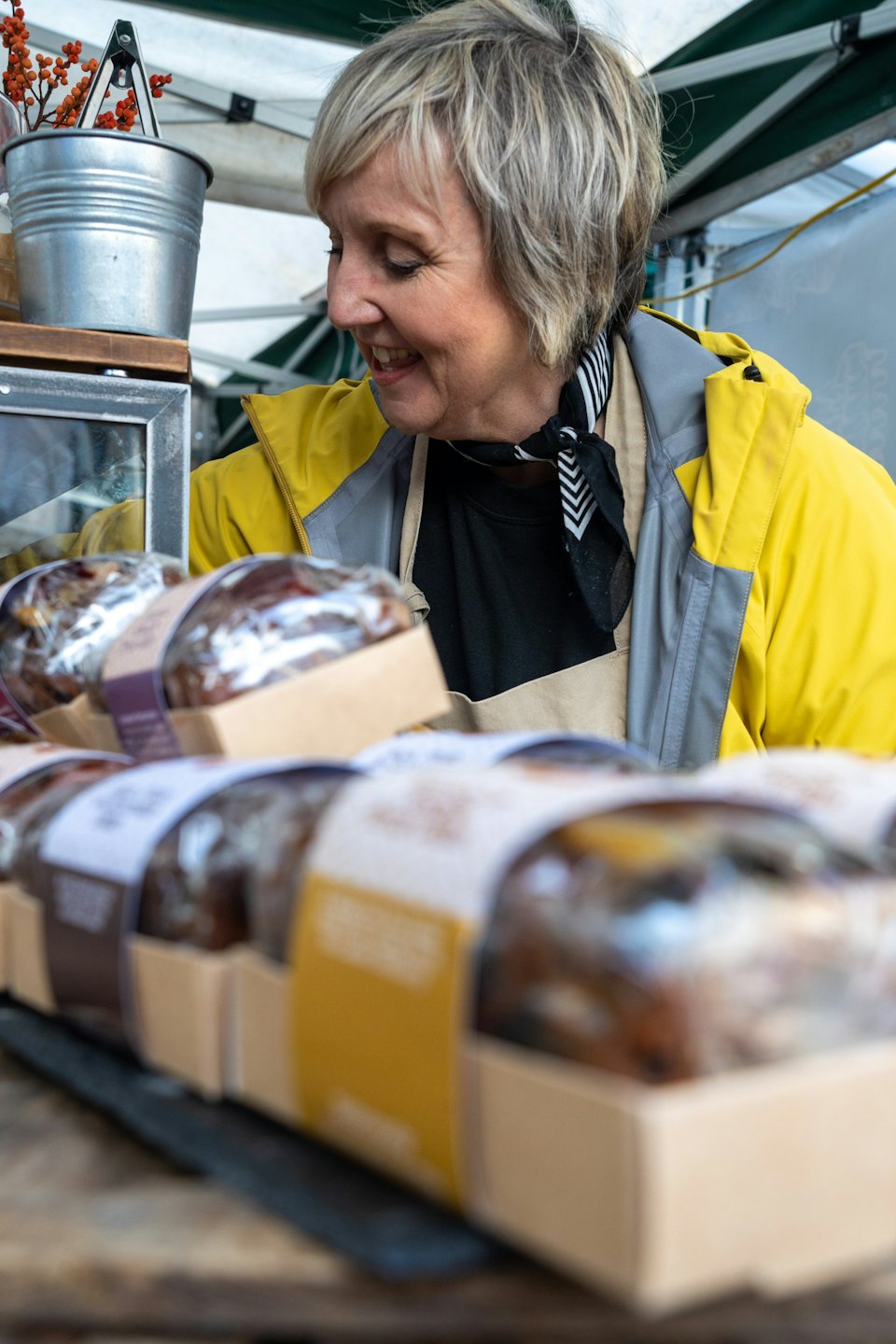 Smiling vendor in yellow jacket looks at baked goods on table.